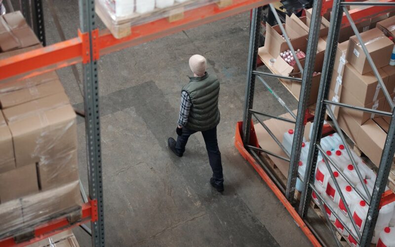 En person går gennem et opbevaringsområde i et lager, omgivet af høje reoler fyldt med papkasser og beholdere.